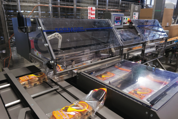 Baked goodies inside a plastic tray are moved through a pair of Formost Fuji horizontal baggers that quickly open up individual plastic bags to allow the trays to enter before sending them on its way to be individually clip-sealed.