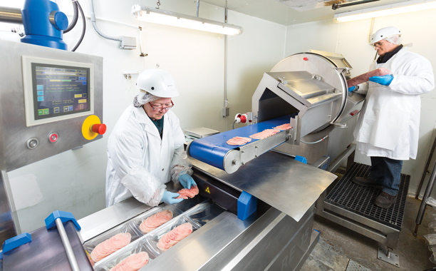Workers place chubs of bison meat through a Holac industrial slicer and then hand-place the fondue slices on the film tray fed to the Repak RE15 form/fill/seal packer.