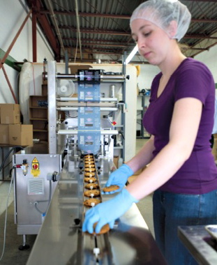 A machine operator manually places freshly-baked cookies onto the infeed conveyor track of the new Sleek 50 Wrapper flowwrapping machine manufactured by Paxiom.